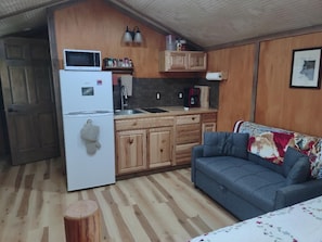 Cocina pequeña equipada con refrigerador, microondas y lavamanos. Hay un armario de madera sobre el lavamanos y un sofá con una manta de motivos florales.