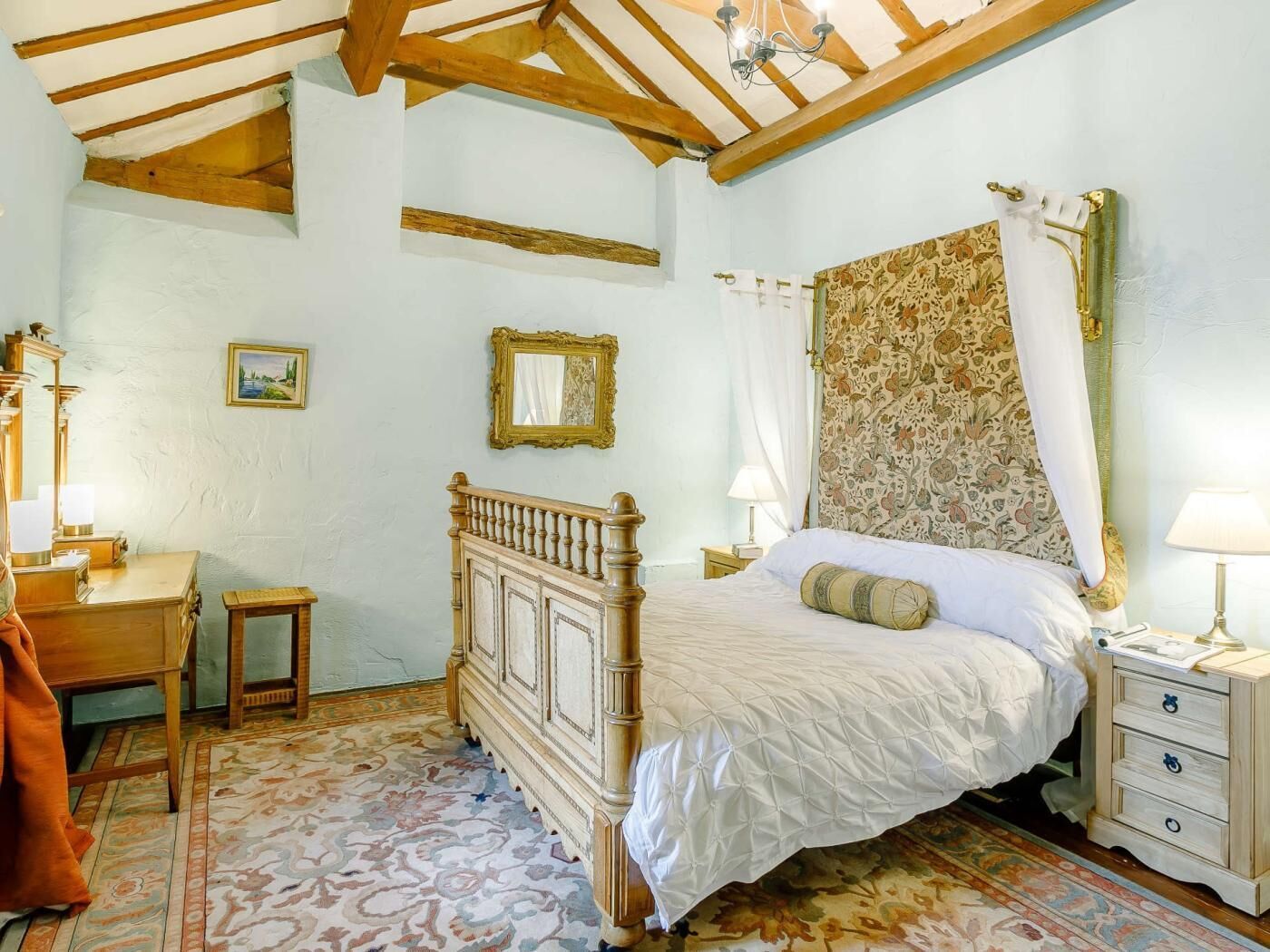 A bedroom with a wooden bed, a desk, a chair, a nightstand, and a patterned carpet.