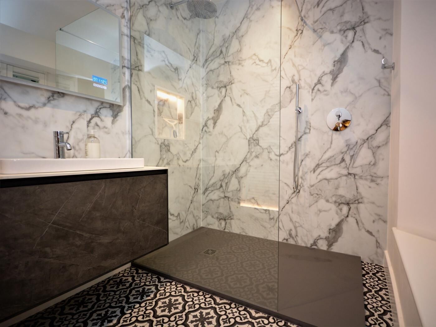Een moderne badkamer met een douchewand van marmer, een glazen douchecabine, een wastafel met een kraan en vloertegels met een patroon.