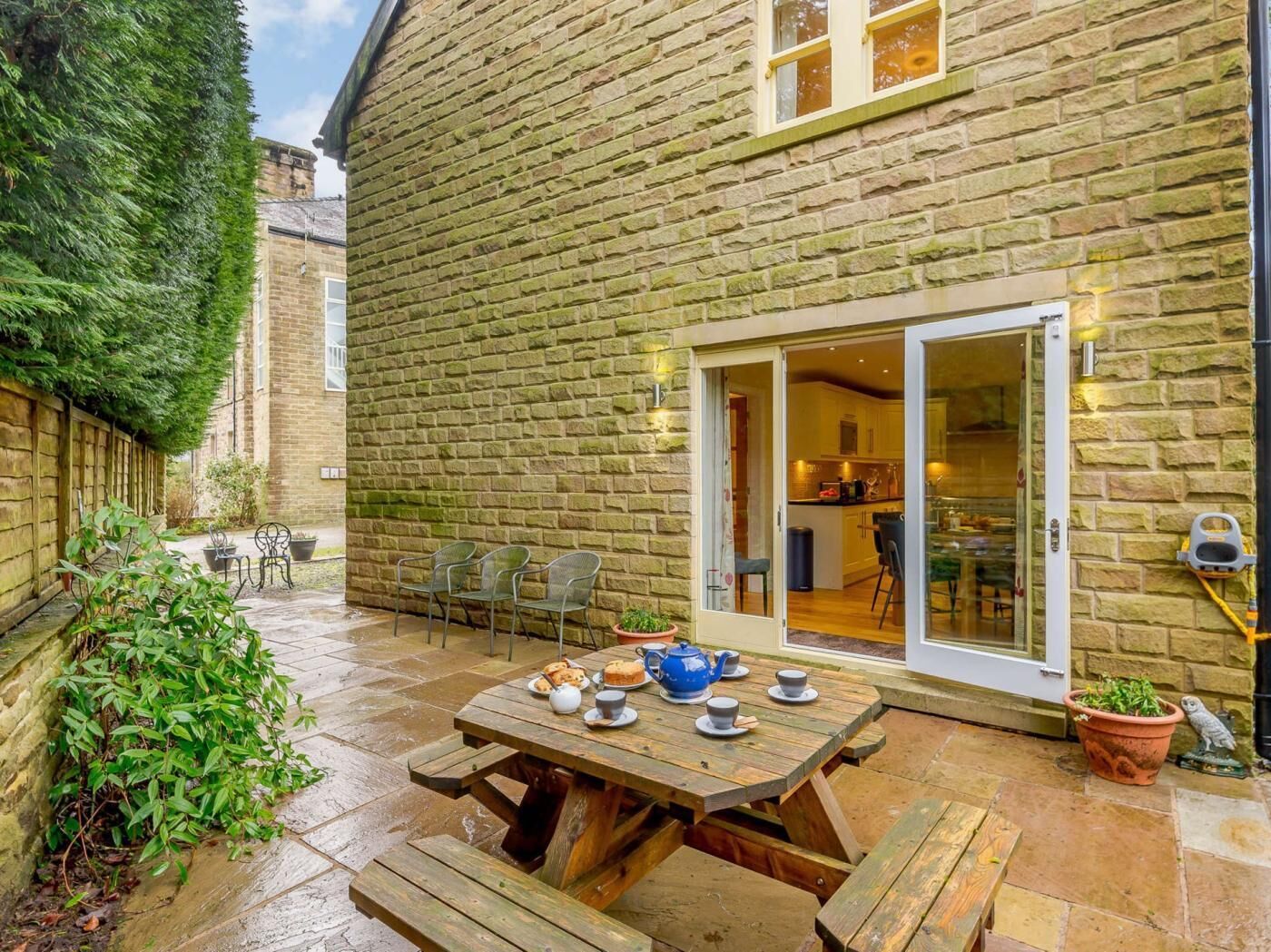 A stone building with a patio area featuring a wooden table set for tea, surrounded by chairs and potted plants.