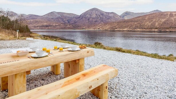 Une table de pique-nique en bois, avec des aliments pour le petit-déjeuner, et une vue sur un lac et des montagnes.