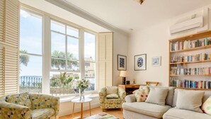 Ein Wohnzimmer mit einer Couch, Sesseln, einem Couchtisch, einem Bücherregal und Blick durch große Fenster.