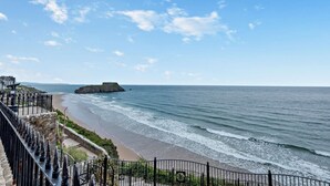 Blick auf die Küste mit einer Steintreppe, die zu einem Strand führt, einer befestigten Struktur auf einer kleinen Insel und einem klaren Himmel.