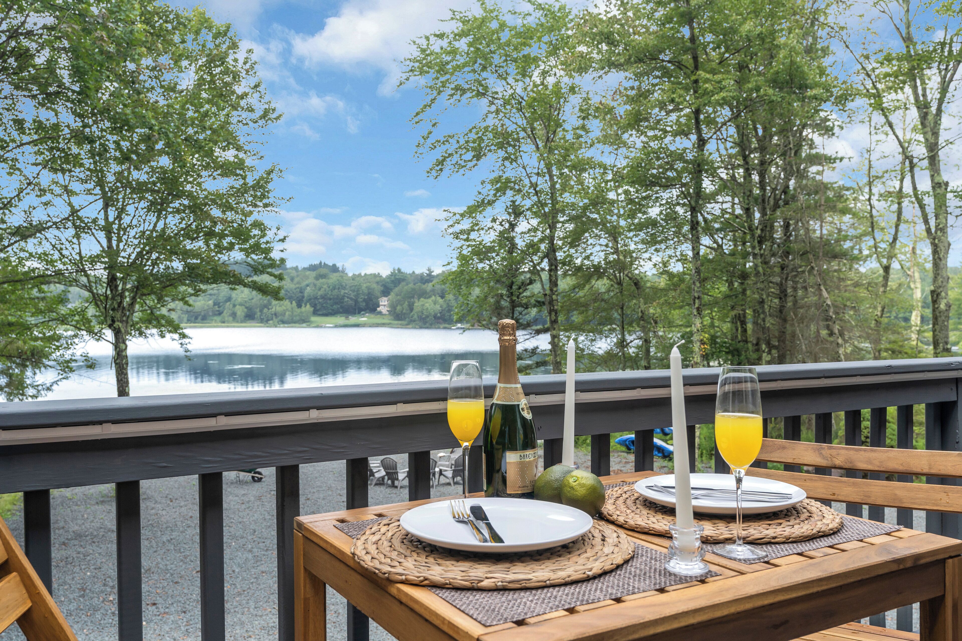 A table set with a bottle, glasses, and a plate, overlooking a lake with trees.