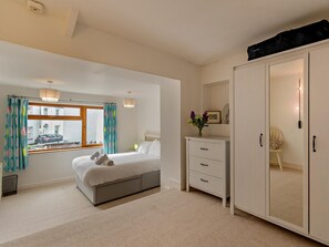 A bedroom with a bed, a dresser, a wardrobe, a window with curtains, and a view of the street outside. - 3 Bed in Tenby (oc-fb231) (Tenby)