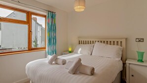 Una cama bien tendida con almohadas y toallas dobladas, una lámpara de noche, una mesita de noche, una ventana con cortinas y un aplique colgante.