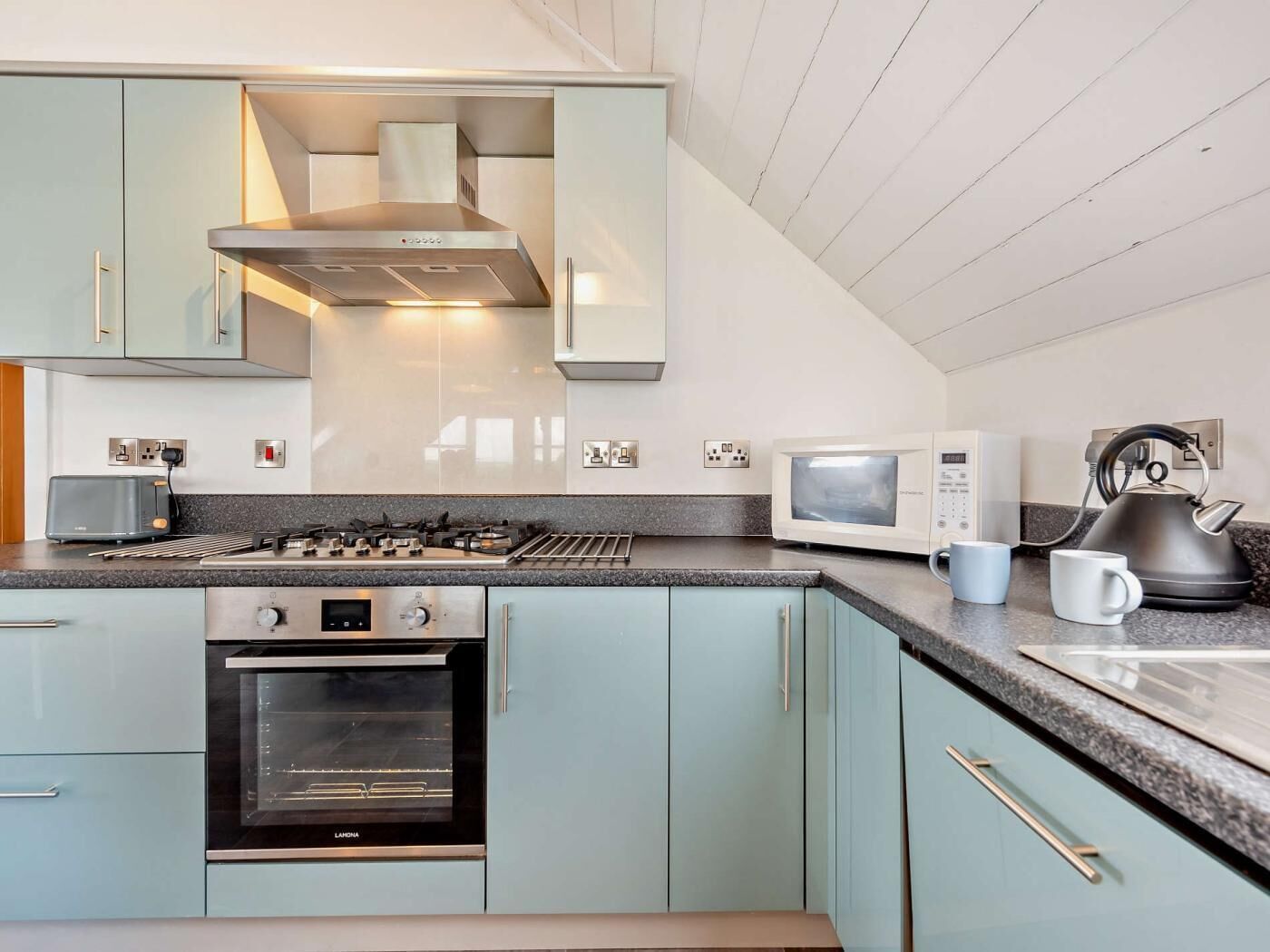 A modern kitchen with light blue cabinets, a stainless steel range hood, a built-in oven, a microwave, and a kettle on the countertop.