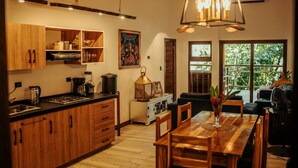 A modern kitchen with wooden cabinets, a dining area with a wooden table and chairs, and a large window with a view of greenery. - Samaya Lush Lakeside Apt - White Lotus (San Miguel)