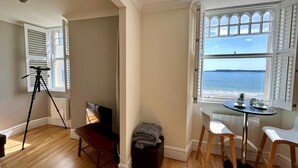 A room with a wooden floor, a tripod, a television, a small table with chairs, and a window with a view of the sea. - 1 Bed in Tenby (oc-fb179) (Tenby)