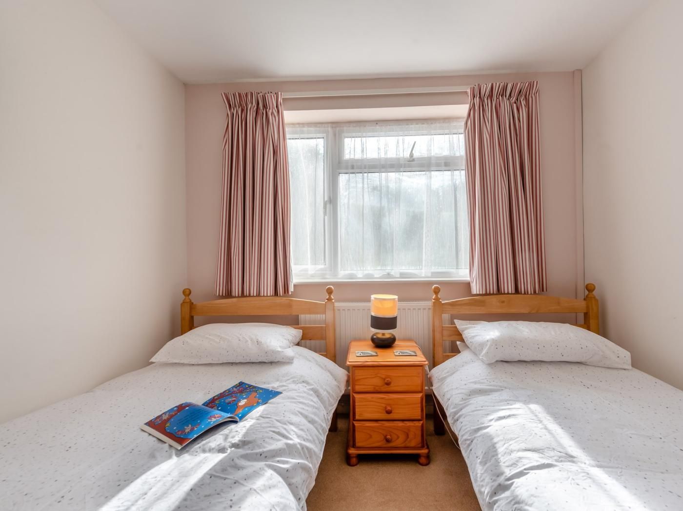 A bedroom with two single beds, a bedside table with a lamp, and a window with curtains.