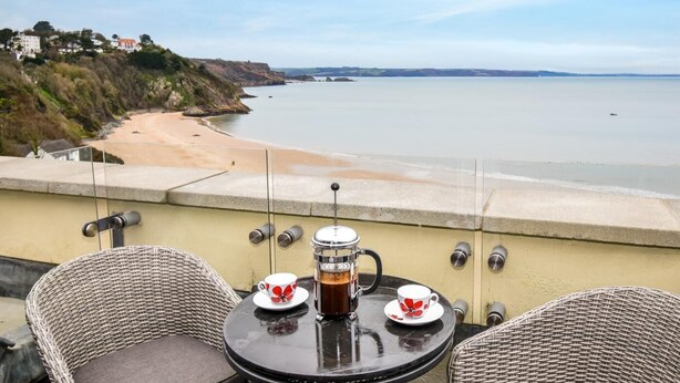 A balcony with wicker chairs, a table with two cups of tea, and a view of the beach and ocean. - 3 Bed in Tenby (oc-fb273) (Tenby)