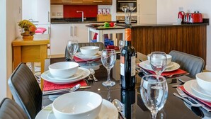 A dining area with a table set for four, a wine bottle, and a kitchen in the background. - 3 Bed in Tenby (oc-fb273) (Tenby)