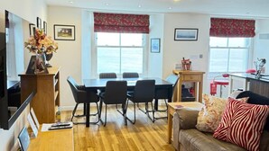 A modern living room with a wooden floor, a sofa with patterned cushions, a dining table with chairs, and a kitchen area with a red chair. - 3 Bed in Tenby (oc-fb273) (Tenby)