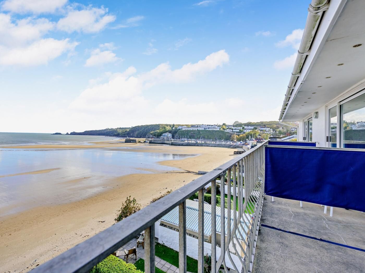 Une vue sur la mer depuis un balcon doté d’une balustrade, une plage de sable et des bâtiments visibles au loin.