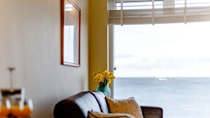 Ein Wohnzimmer mit einer Couch, einem Blumenstrauß aus gelben Blüten und Meerblick durch ein Fenster.