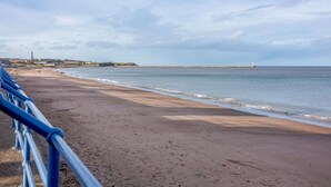 Ein Strand mit Metallgeländer, ein Leuchtturm in der Ferne und ein klarer Himmel.