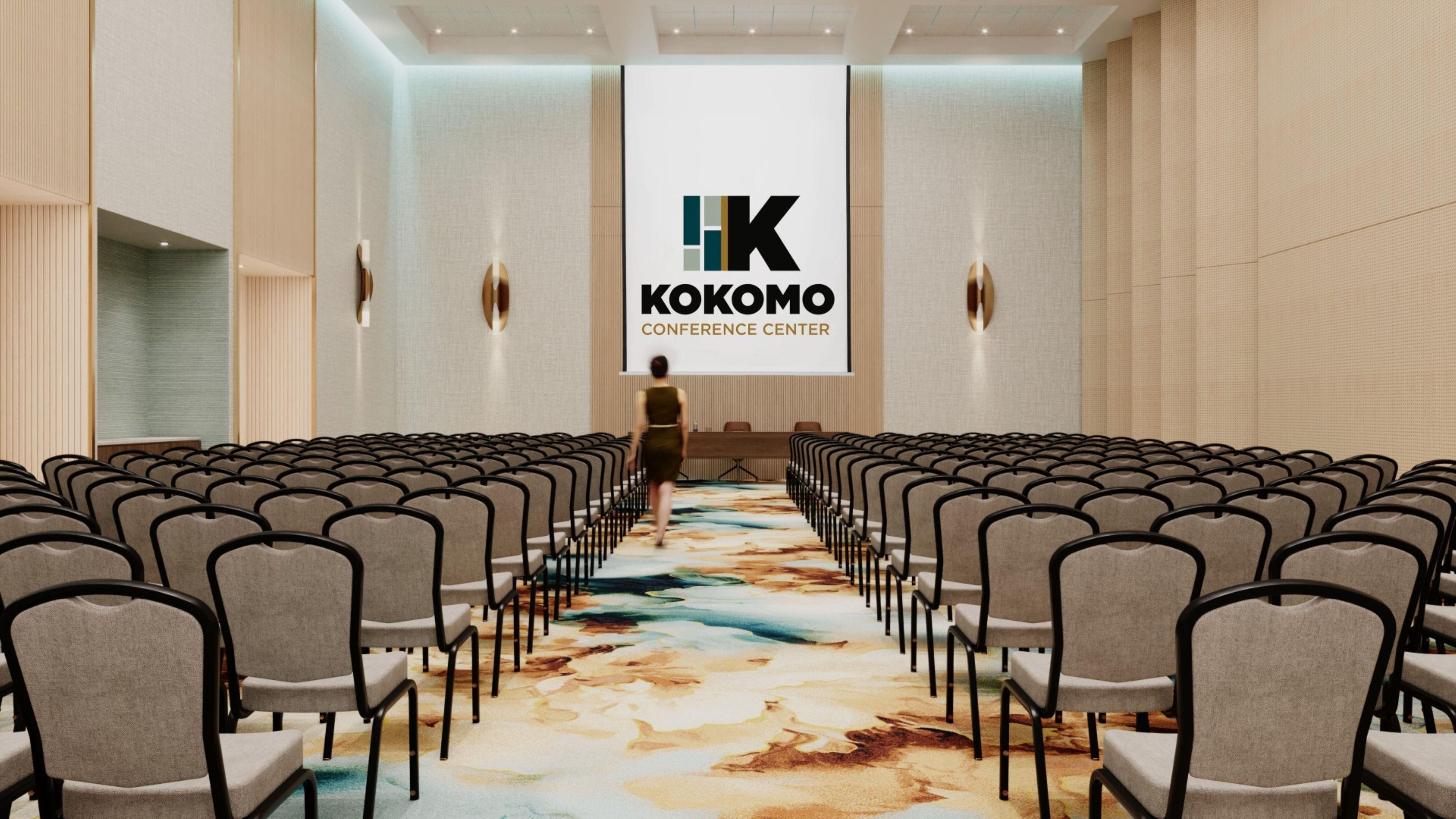 Un salón de conferencias con filas de sillas mirando hacia un escenario, una pantalla grande que muestra 'KOKOMO CONFERENCE CENTER' y una alfombra estampada.