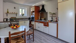 Een keuken met een eettafel, stoelen, een mand met fruit, een fornuis, een spoelbak en een koelkast.