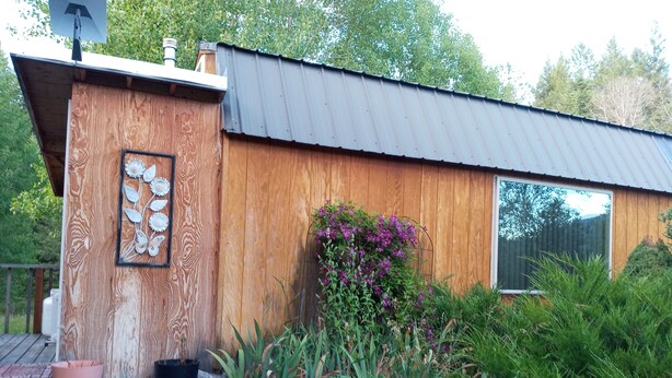 A wooden house with a metal roof, a window, and a garden with plants and a butterfly decoration. - Cozy one bedroom tiny house. (Colville)