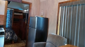 A room with a wooden table, a refrigerator, a chair, a clock on the wall, and a window with blinds. - Cozy one bedroom tiny house. (Colville)