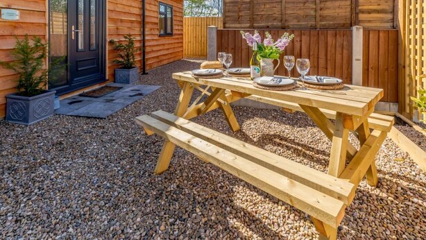Une cabane en bois avec un patio en gravier, une table à manger dressée avec des assiettes, des verres et un vase de fleurs.