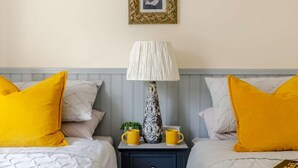 Une chambre d’hôtel avec deux lits, chacun recouvert d’une couette blanche et agrémenté d’oreillers jaunes, une table de chevet avec une lampe, un tableau encadré au mur et une petite plante posée sur la table de chevet.