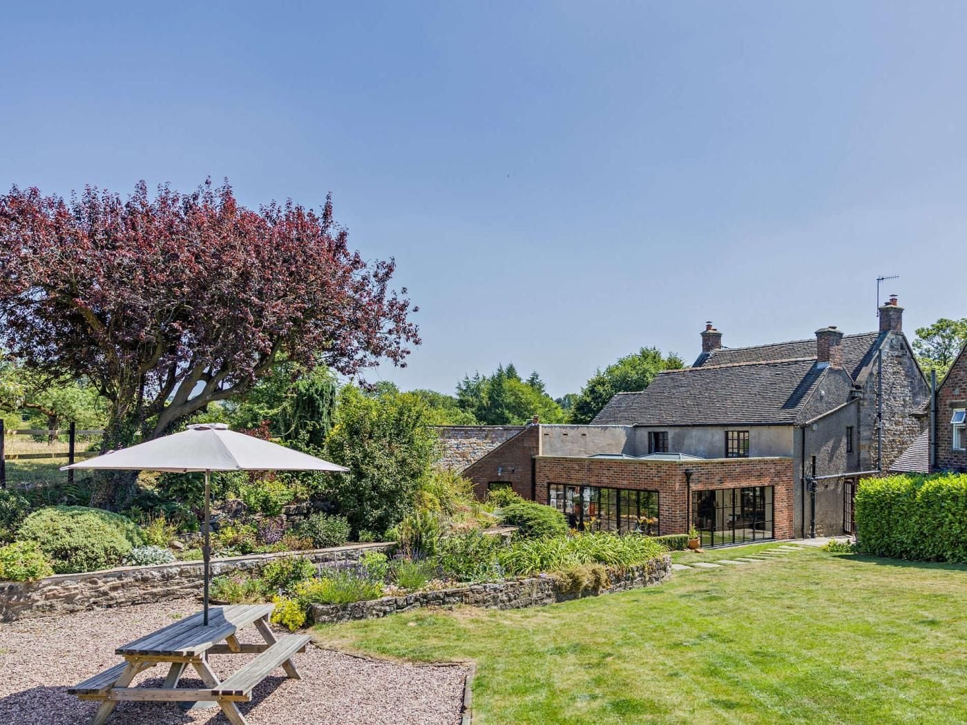 A traditional stone house with a large garden, a picnic table, and a tree with red leaves.