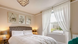 Een slaapkamer met een groot bed, twee nachtkastjes, een kroonluchter, een raam met gordijnen en kunstwerken aan de muur.