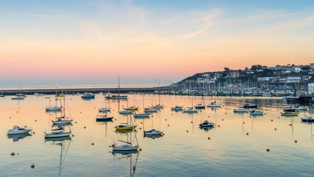 Een jachthaven met aangemeerde boten, rustig water en een stad op de achtergrond.