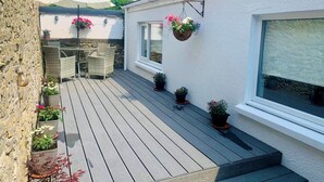 A wooden deck with outdoor furniture, potted plants, and hanging flower baskets. - 2 Bed in Tenby (oc-fb261) (Tenby)