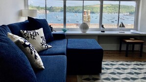 Un salon avec un canapé bleu, une petite table, un vase et une vue sur des bateaux sur l’eau.