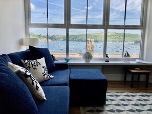 Ein Wohnzimmer mit einem blauen Sofa, einem kleinen Tisch, einer Vase und Blick auf Boote auf dem Wasser.
