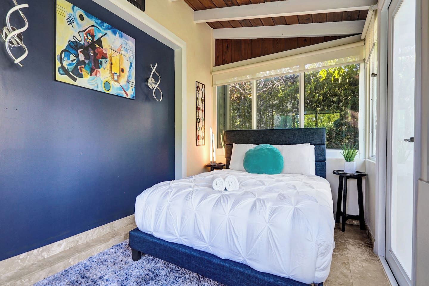 A bedroom with a bed, a nightstand, a painting, and a window with a view of trees.
