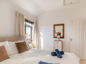 Ein Schlafzimmer mit einem Bett, einem Nachttisch mit einer Vase, einem Spiegel und einem Fenster mit Vorhängen.