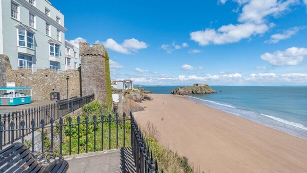 A coastal view with a sandy beach, a stone wall, and a building with multiple windows. - 2 Bed in Tenby (oc-fb233) (Tenby)