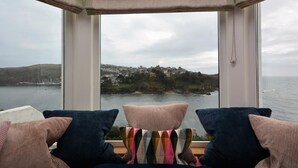 A room with a large window overlooking a coastal view, featuring a chair with a colorful cushion and two dark pillows. - 3 Bed in Fowey (oc-81112) (Fowey)