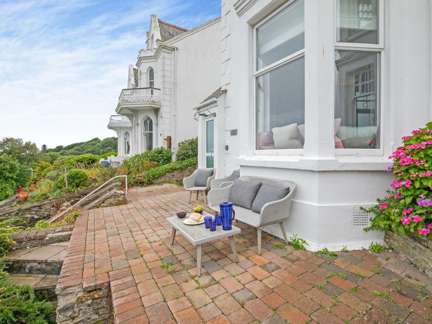 Un bâtiment blanc doté d’une baie vitrée, un petit espace extérieur aménagé avec une table et des chaises, et un jardin avec différentes plantes et fleurs.