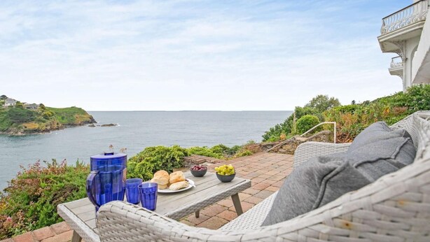 A wicker chair with a cushion, a table with food and drinks, and a view of the sea. - 3 Bed in Fowey (oc-81112) (Fowey)