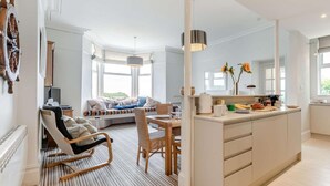 A modern kitchen with a central island, a dining area with a wooden table and chairs, and a seating area with a sofa and a rocking chair. - 3 Bed in Fowey (oc-81112) (Fowey)