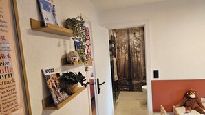 Et værelse med en træhylle med bøger og planter, en bamse på et bord, og en dør der fører til et badeværelse med skovtapet.