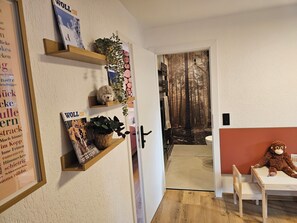 Ein Zimmer mit einem hölzernen Regal, das Bücher und Pflanzen beherbergt, einem Teddybären auf einem Tisch und einer Tür zu einem Badezimmer mit Waldtapete.