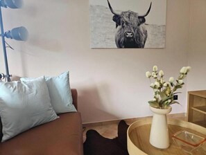 Ein Wohnzimmer mit einem braunen Sofa, einer weißen Vase mit Blumen, einer Lampe und einem gerahmten Bild an der Wand.