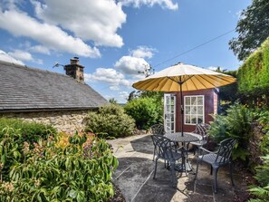 Une maison en pierre avec un patio carrelé, une table ronde entourée de chaises et un grand parasol.