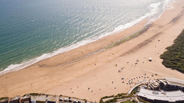 An aerial view of a beach with a curved structure, likely a hotel or resort, with a clear view of the ocean and distant landforms. - 2 Bed in Tenby (oc-fb285) (Tenby)