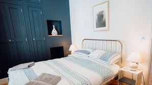 A neatly made bed with striped bedding, a bedside table with a lamp, and a wall-mounted shelf with a decorative item. - 2 Bed in Tenby (oc-fb285) (Tenby)
