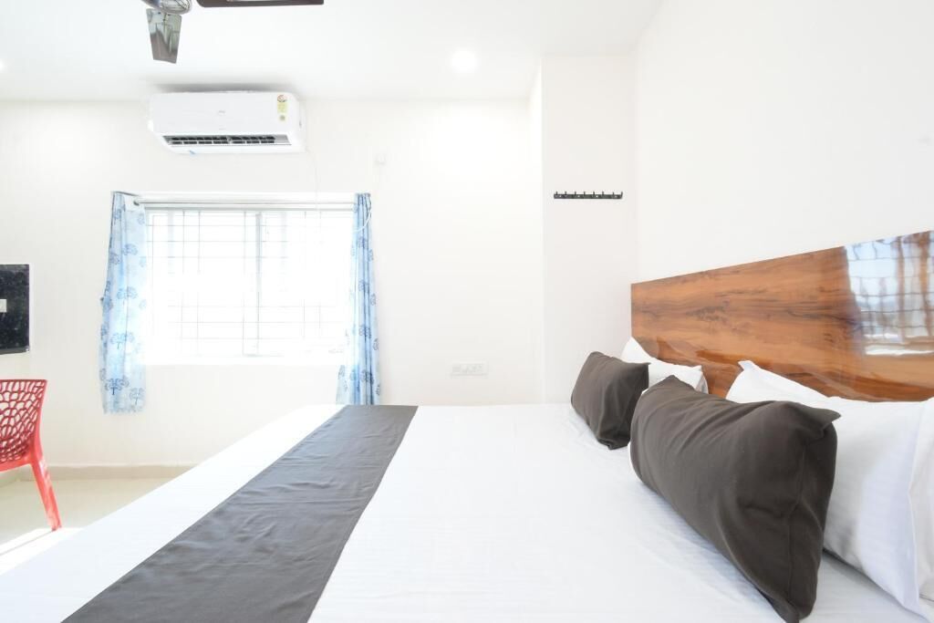 A hotel room with a bed, a red chair, an air conditioning unit, a window with curtains, and a wooden headboard.
