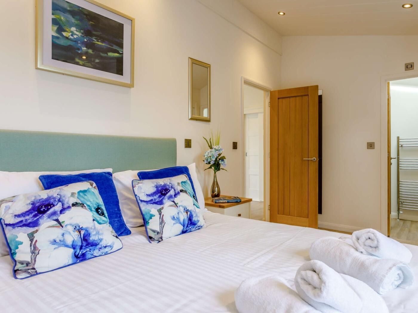A hotel room with a bed, pillows, a vase with flowers, a wooden door, and a towel rack.