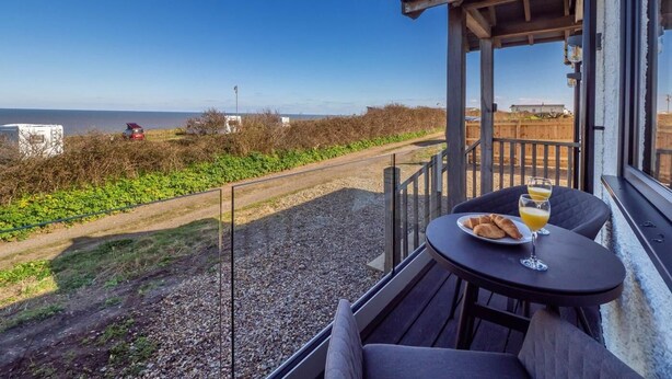 A balcony with a table set for two, overlooking a coastal road and the sea. - 1 Bed in Norwich (oc-82230) (Mundesley)