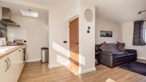 A modern kitchen with wooden flooring, a dining area with a dark sofa, and a view of the outdoors. - 1 Bed in Norwich (oc-82230) (Mundesley)
