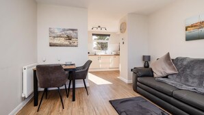 A modern living room with a sofa, a dining table with chairs, a kitchen in the background, and a painting on the wall. - 1 Bed in Norwich (oc-82230) (Mundesley)